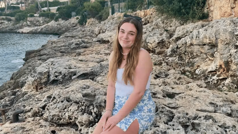 A young white women sat on rocks by a beach.